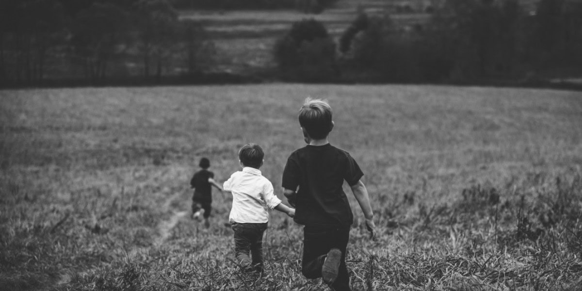 Tres niños corren por un campo abierto, dos en primer plano y uno más atrás. La escena es en blanco y negro, evocando un sentimiento temporal de celos entre hermanos que sabemos que pronto desaparecerá mientras juegan bajo los árboles distantes.