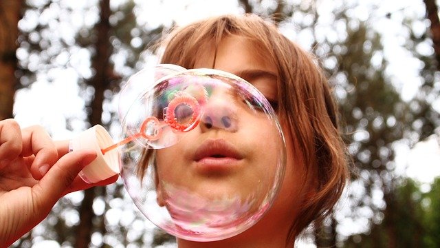 Un niño practicando motricidad orofacial mientras sopla alegremente una burbuja al aire libre, con árboles que enmarcan maravillosamente el fondo.