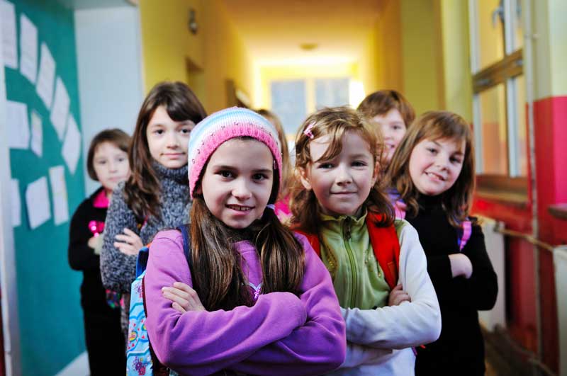 Un grupo de niños pequeños se encuentra en el pasillo de una escuela, algunos se cruzan de brazos y otros sonríen. Vestidos con ropa de invierno, disfrutan de las vibrantes paredes del pasillo mientras aprenden a mejorar habilidades sociales y construir amistades duraderas.