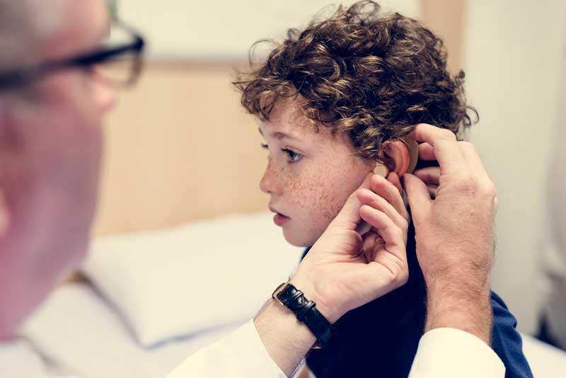 Un niño con cabello rizado recibe ayuda de un adulto ajustando un audífono en un entorno clínico, abordando los desafíos de la comunicación en sordoceguera.