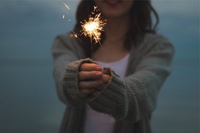 Una persona con un suéter gris sostiene una bengala encendida con ambas manos contra un fondo tenuemente iluminado, celebrando el nuevo año y la promesa de nuevos objetivos.