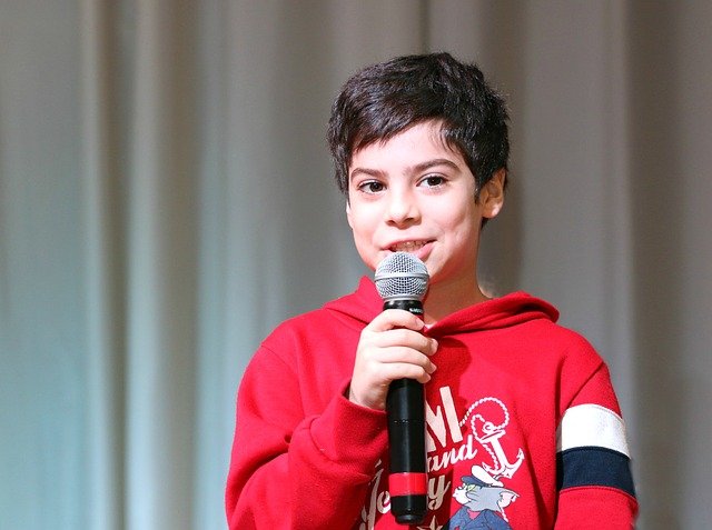 Un niño pequeño con una sudadera roja está en el escenario sosteniendo un micrófono, sonriendo y encarnando el espíritu del aprendizaje.