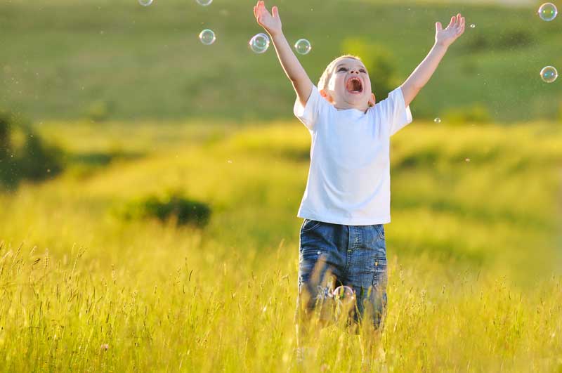 Un niño pequeño con TDAH vestido con una camisa blanca y jeans se encuentra en un campo con los brazos levantados, sonriendo con entusiasmo ante las burbujas que flotan a su alrededor.