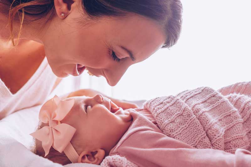 Una mujer sonríe y se inclina hacia un bebé que también sonríe. El bebé, envuelto en una manta rosa y luciendo una diadema con lazo rosa, se beneficia de esta atención temprana, poniendo de relieve la importancia de la neuropsicología para favorecer un desarrollo saludable.