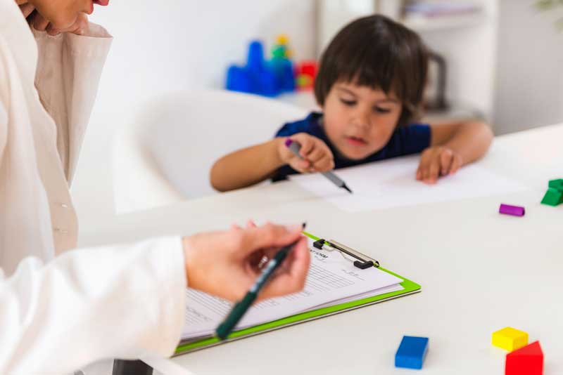 Un niño está sentado a una mesa dibujando con un bolígrafo. Un adulto cercano sostiene un portapapeles y un bolígrafo, observa al niño y posiblemente toma nota de factores para la terapia. Hay bloques de colores esparcidos sobre la mesa.