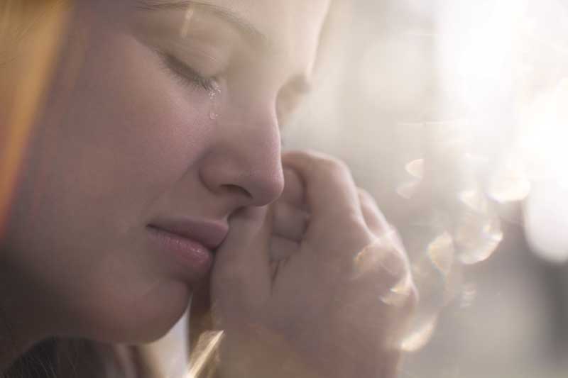 Una imagen en primer plano del rostro de una persona con los ojos cerrados y una lágrima rodando por su mejilla, su mano apoyada cerca de su boca, bañada por una luz suave y difusa que amplifica sutilmente el sentimiento de tristeza.