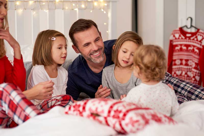 Un hombre y tres niños están sentados juntos en una cama en una habitación decorada con luces navideñas. Están sonriendo, vistiendo pijamas navideños y disfrutando del espíritu festivo de la temporada de fiestas.