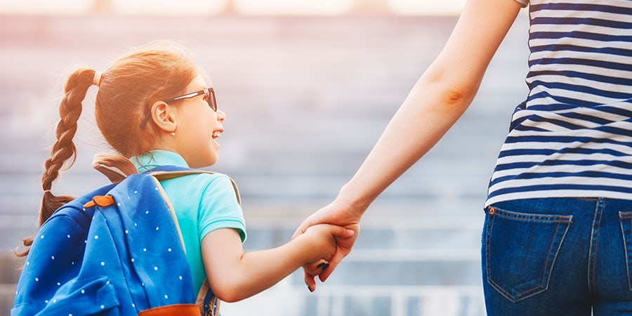 Un niño con gafas de sol y una mochila toma de la mano a un adulto, caminando al aire libre en un día soleado. Es un momento que marca el final de PEREZA y el comienzo de LA VUELTA A LA RUTINA, mostrando cómo estas rutinas necesarias dan forma a nuestras vidas.