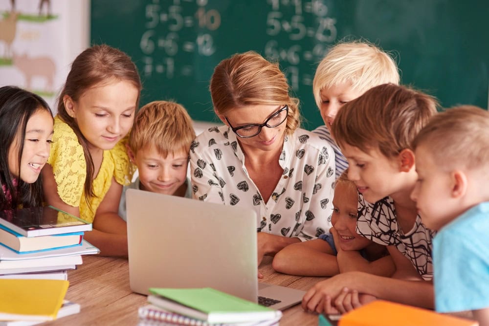 Una maestra y un grupo de niños se reúnen alrededor de una computadora portátil en un salón de clases, promoviendo Disciplina Positiva, con libros y una pizarra de fondo.