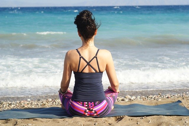 Una persona sentada con las piernas cruzadas sobre una estera de playa frente al océano, vestida con una camiseta sin mangas oscura y mallas coloridas, con el cabello recogido. Inmersos en la meditación, disfrutan de los beneficios del bienestar mientras las olas se acercan suavemente a la orilla al fondo.