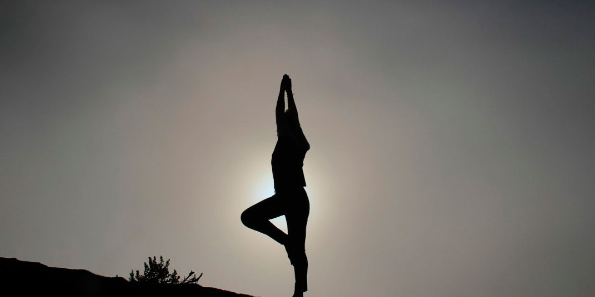 Silueta de una persona realizando una postura de árbol en yoga, de pie sobre una pierna con las manos levantadas por encima de la cabeza contra el fondo de un cielo poco iluminado, encarnando la herramienta de autosuperación.